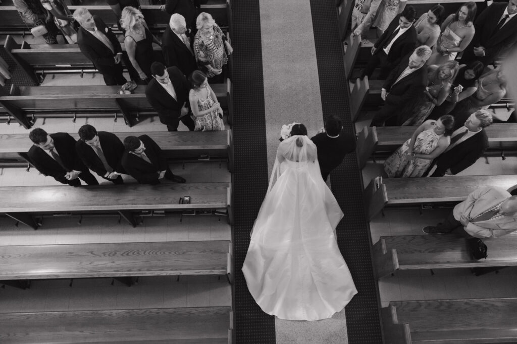 Black and white documentary-style photo of the father of the bride and the bride walking down the church aisle during the wedding ceremony.
