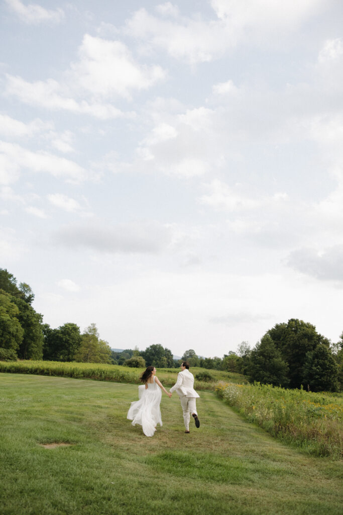 A newlywed couple in their wedding dress and suit joyfully runs through a green field of an upstate New York wedding venue. Documentary-style wedding photography captures intimate, motion imagery of the bride and groom. 