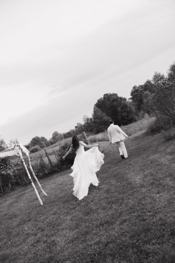 The bride and groom of an upstate New York intimate, luxury wedding run across a field, holding hands. Documentary wedding photography captures real-time romance, freedom, and intimacy. 