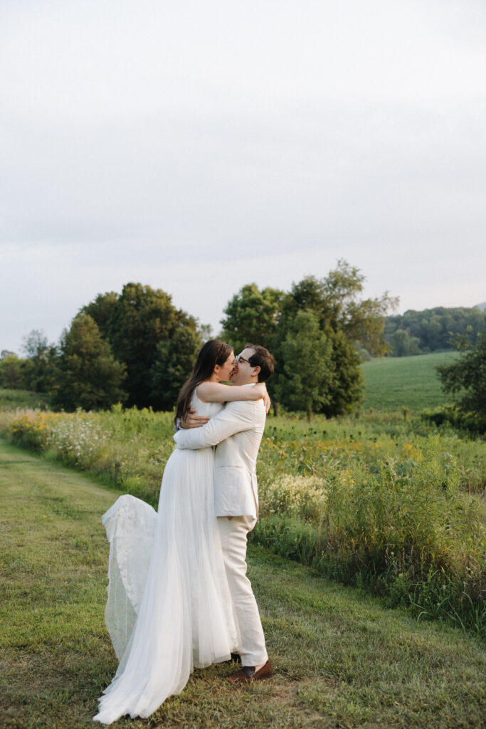 A newlywed couple embraces lovingly in a lush, green field of an upstate New York wedding venue. Intimate wedding photography creates serene, romantic, and emotional imagery.