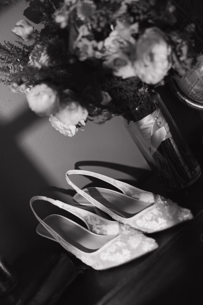 Black and white documentary-style wedding photography, elegant wedding high-heeled shoes with lace detail next to a vase of flowers. Wedding photos convey timeless elegance and anticipation of a Hudson Valley, New York wedding.