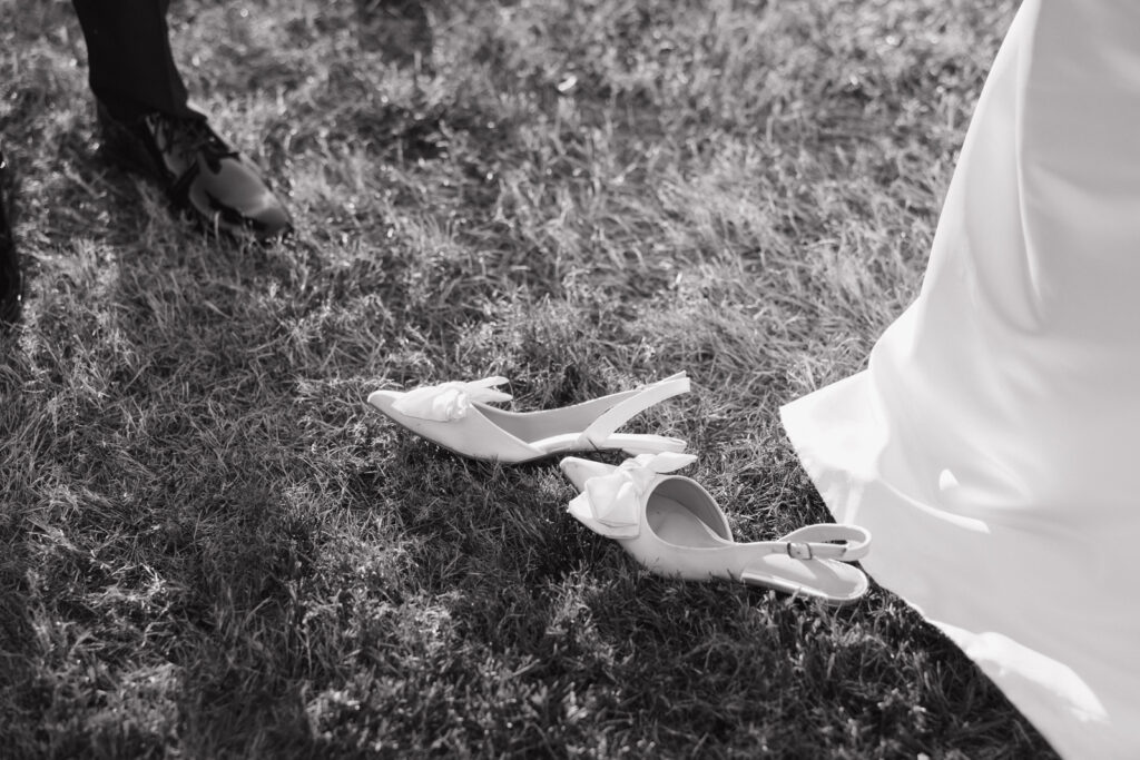 Black and white documentary style wedding photography. The brides white heels with a bow on the toe are laying in the grass next to the brides dress.