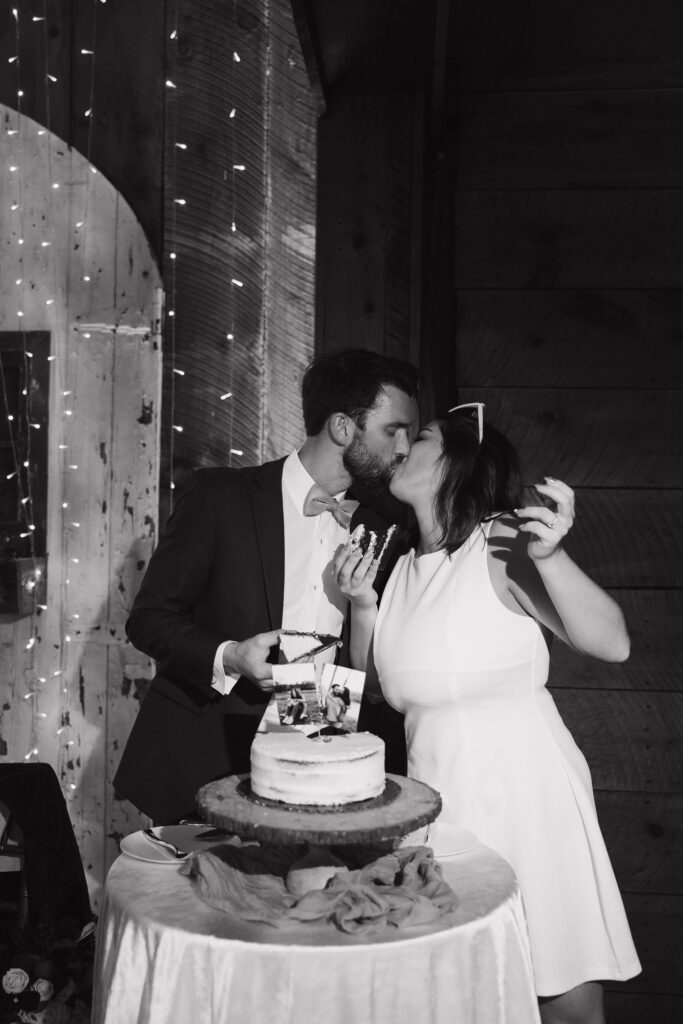 Black and white documentary-style photography capturing newlyweds sharing a kiss after cutting their wedding cake.