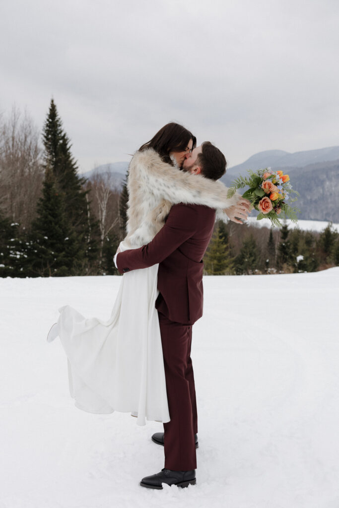 The groom is holding the bride with the Adirondack Mountain range in the background, covered in snow. 