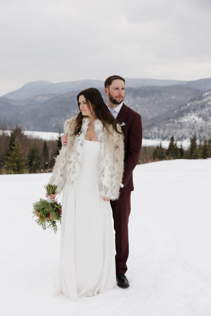 Documentary style wedding photographer captures the bride and groom looking out, while the snow-covered Adirondack Mountain range spans the background behind the couple.