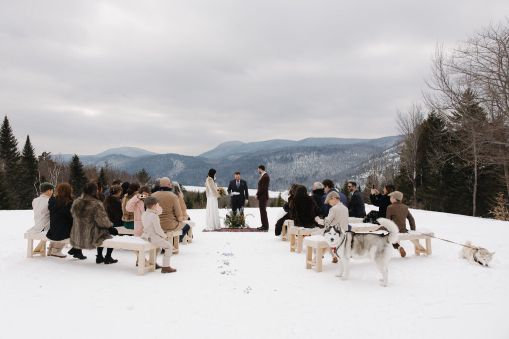 The bride and groom stand facing each other during their outdoor ceremony at their Adirondack winter wedding. Small, intimate wedding outside, with guests sitting on each side of the aisle and the Adirondack mountain range in the background.