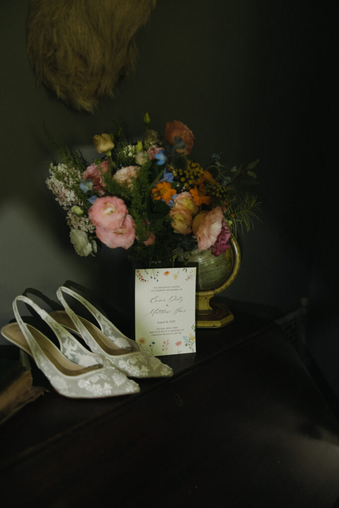 The bride's beautiful pastel-colored bouquet, wedding invitation, and white heels are propped with sunlight beaming onto them.