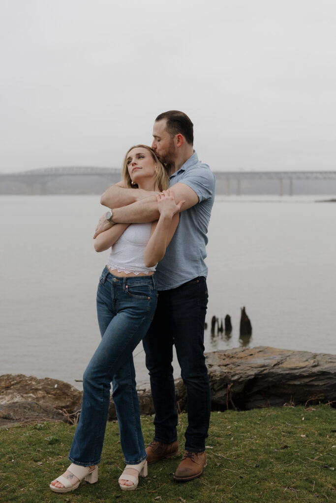 Couple embracing by the water during a romantic Guilford Connecticut engagement session with a soft coastal backdrop.