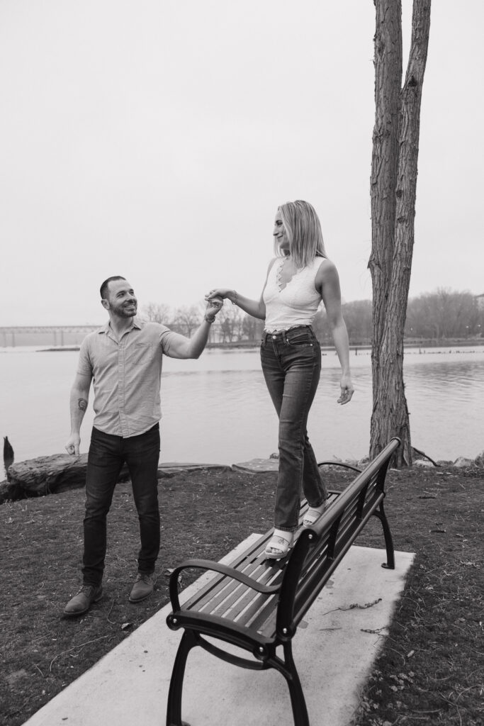 Couple laughing and walking hand-in-hand by the water during a Connecticut engagement session with a soft, overcast sky.