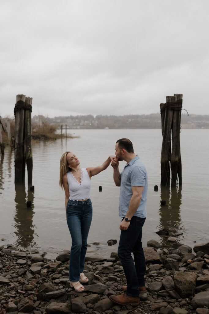 Couple romantically posed by the water during an engagement session with a soft, overcast sky.