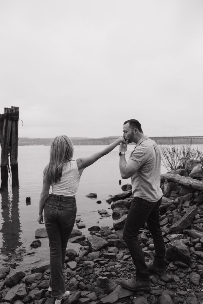 Black and white photo of a couple sharing a kiss on the hand while walking along the water