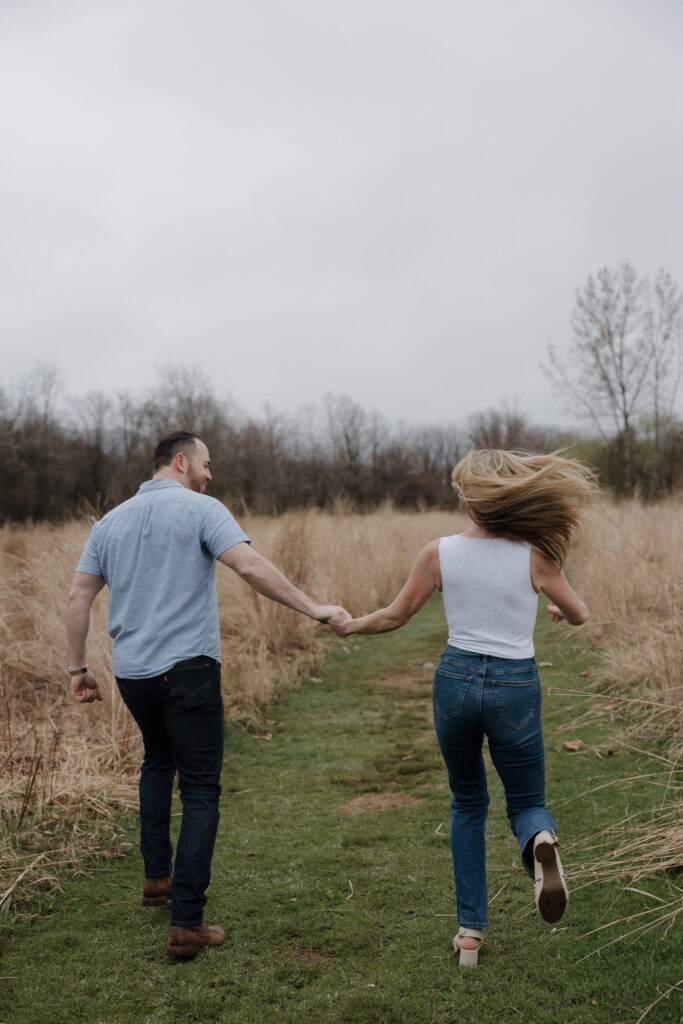 Couple running hand in hand through a grassy field during an engagement session.