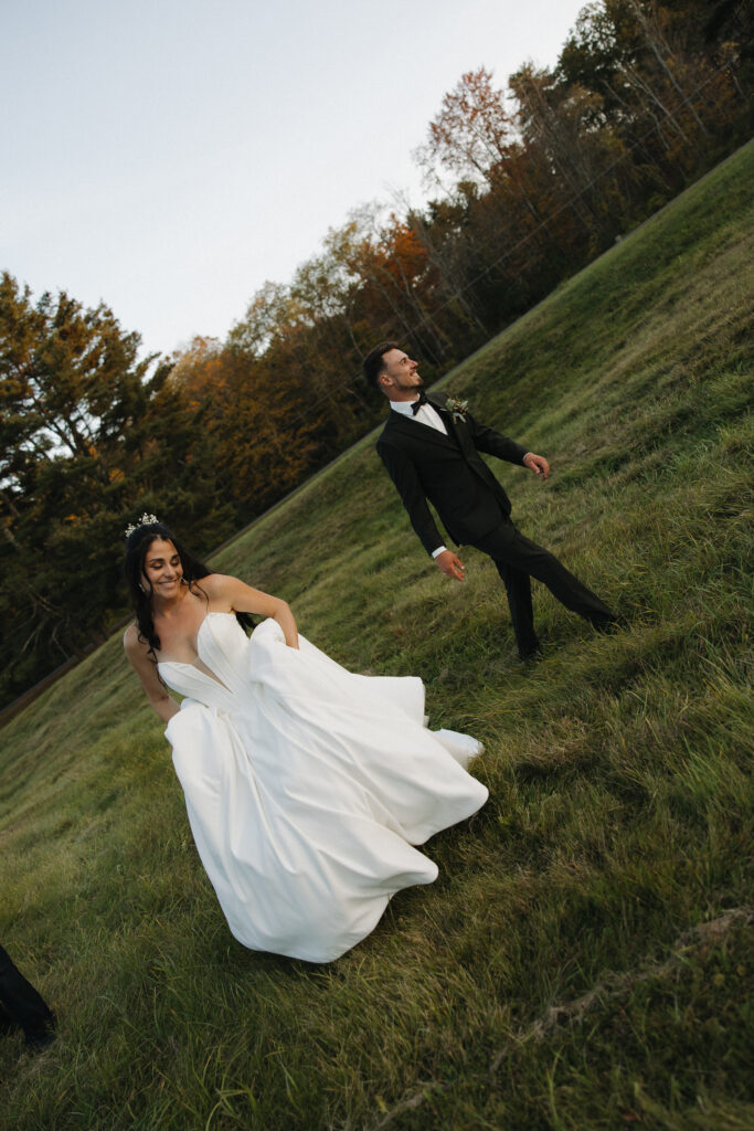 Bride and groom laughing and running together through a green hillside field surrounded by fall foliage during wedding portraits