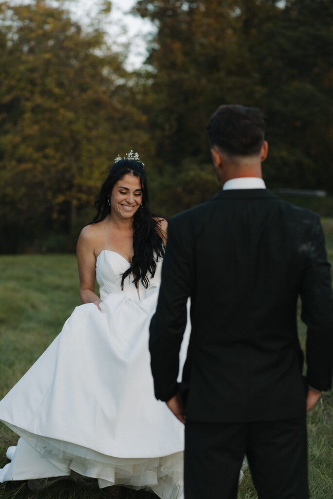 Bride in a strapless white ball gown smiling and twirling while groom looks on during outdoor wedding portraits in a green field
