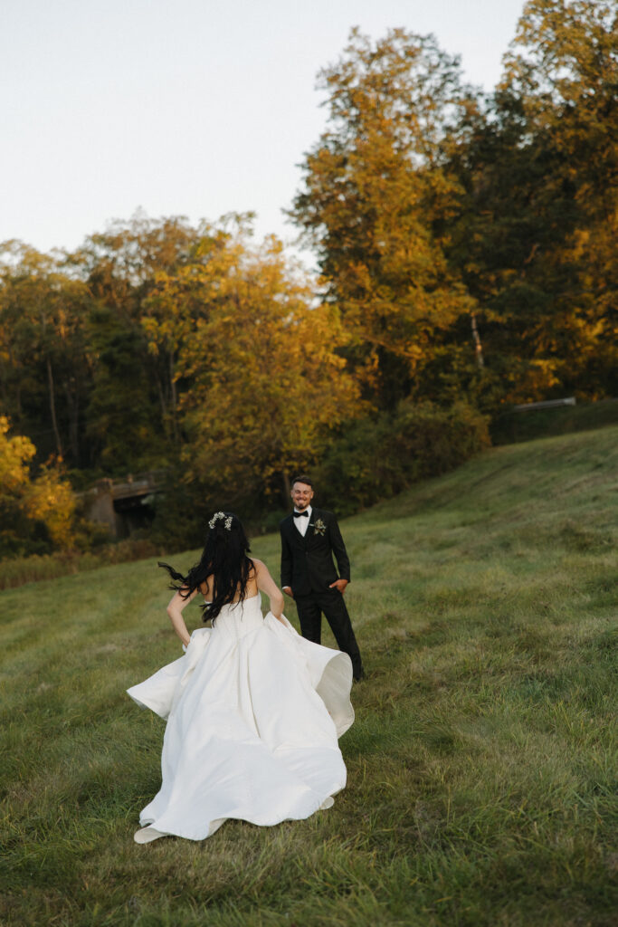  Bride in a flowing white gown running toward groom on a grassy hill with golden autumn trees in the background during sunset portraits