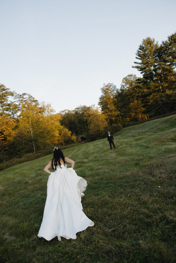  Bride in a white ball gown walking across a wide open grassy field toward groom standing at a distance surrounded by fall trees
