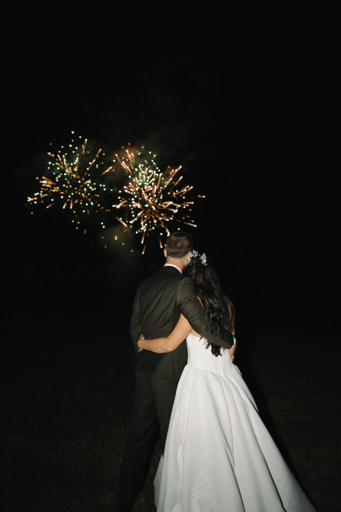  Bride and groom holding each other from behind watching colorful fireworks burst in the night sky at their wedding