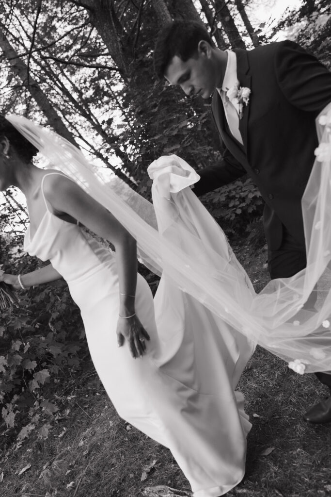 black and white documentary style photo of groom carrying brides dress while walking 