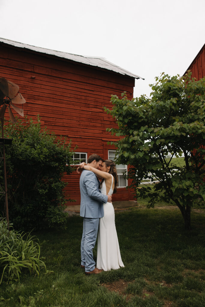 Couple share an intimate moment in front of a rustic red barn, groom in a light blue suit and bride in a backless white wedding dress