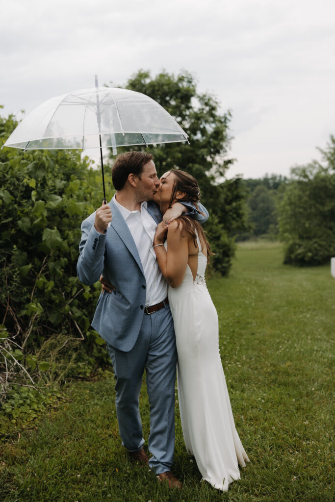 Bride and groom kiss under a clear umbrella on a rainy wedding day in a green Connecticut field